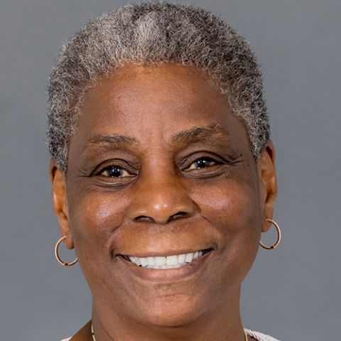 A woman with very short salt and pepper hair smiles in front of a gray studio backdrop