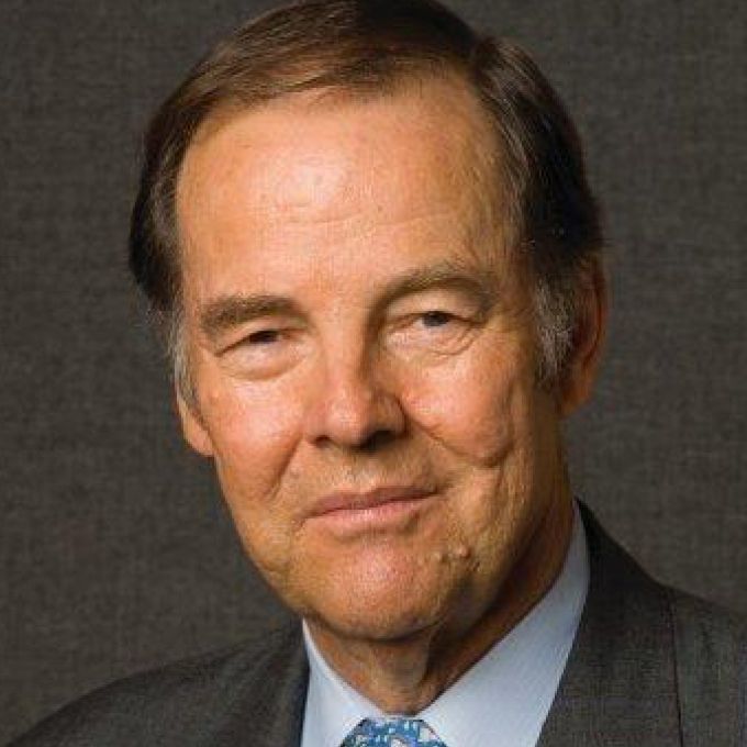 A man smiles in front of a dark studio background in gray jacket, light blue shirt, and patterned tie