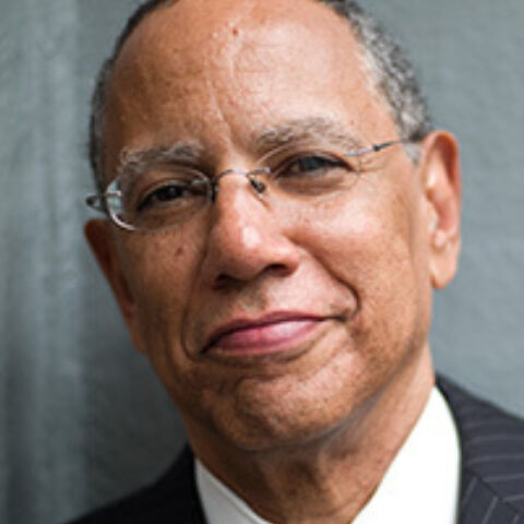 A man smiles in front of a gray wall in frameless eyeglasses and dark jacket