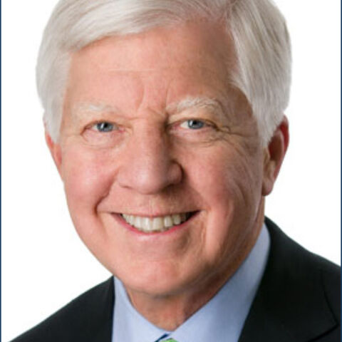 Man with gray hair smiling in a dark jacket, light blue dress shirt, and green patterned tie