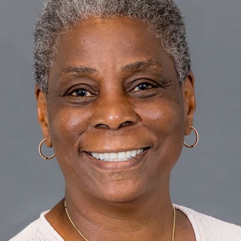 A woman with very short salt and pepper hair smiles in front of a gray studio backdrop