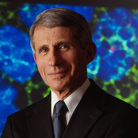 A man poses in front of an abstract blue and green cellular background in a dark jacket, white shirt, and blue tie