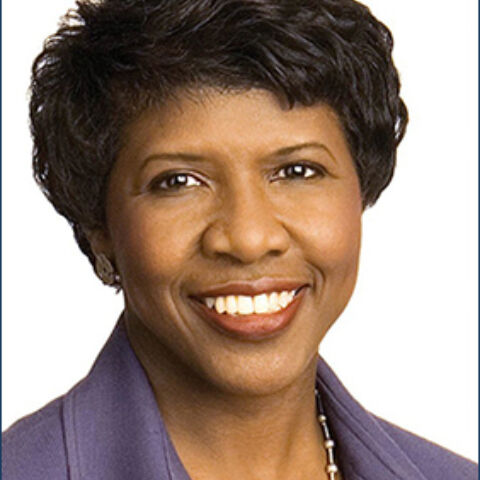 A woman with black hair smiles warmly in a purple blouse with a sliver necklace