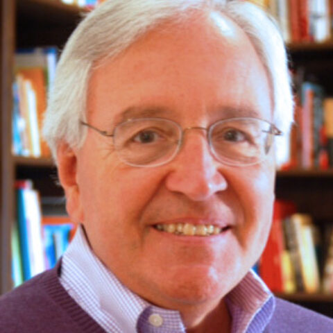 A man with gray hair and rimless glasses poses in front of a bookshelf in a purple sweater and dress shirt