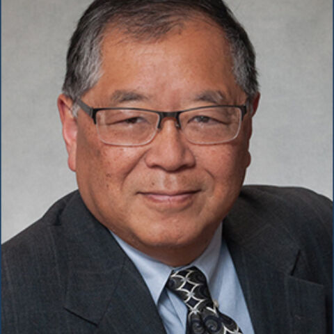 A man with graying hair and square-rim glasses poses in a dark jacket, light blue dress shirt, and patterned tie