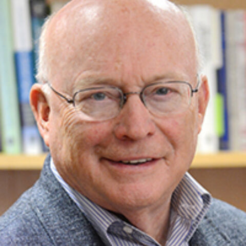 A man poses in front of a bookshelf in a gray jacket and checked shirt