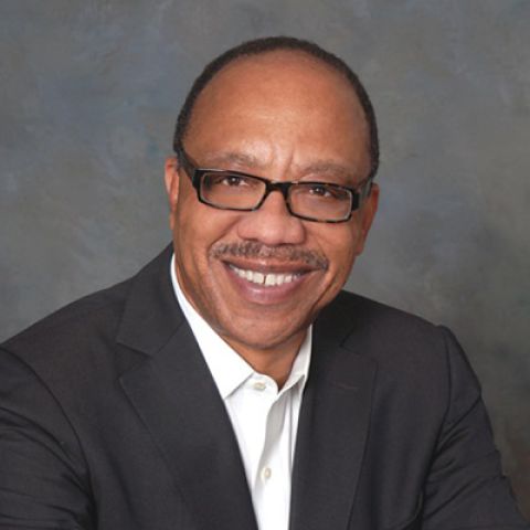 A man smiles broadly while posing in front of a gray studio background in a dark jacket and white dress shirt