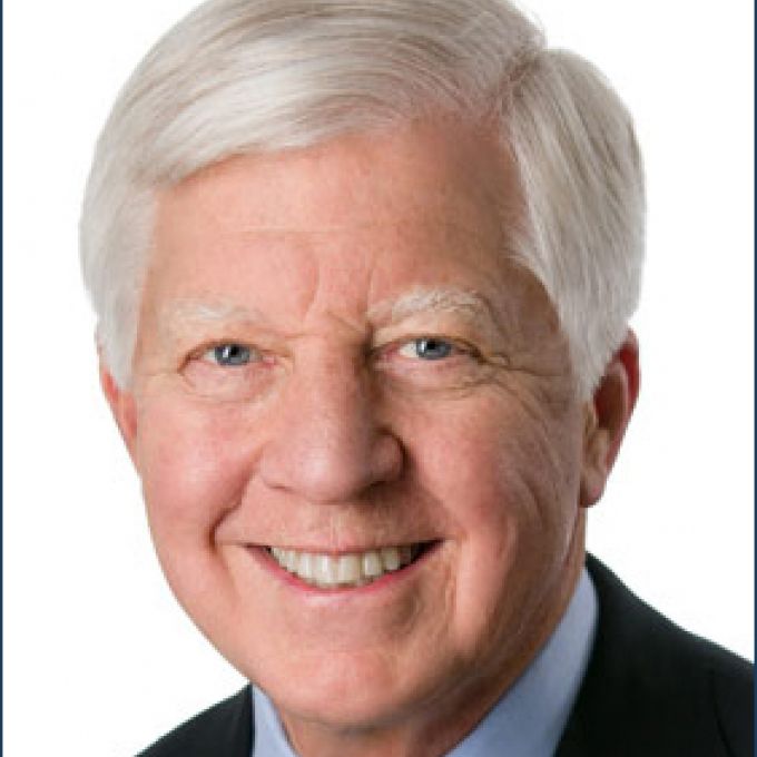 Man with gray hair smiling in a dark jacket, light blue dress shirt, and green patterned tie