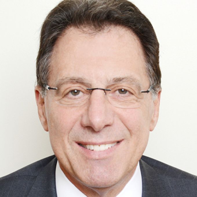 A man with neat brown hair poses in rimless eyeglasses wearing a dress jacket, white white, and light blue tie