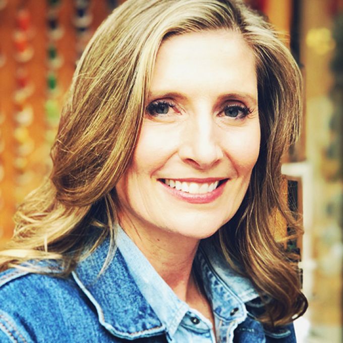 A woman with shoulder-length light brown hair smiles broadly in front of a rust-colored backdrop in a jean jacket and denim shirt