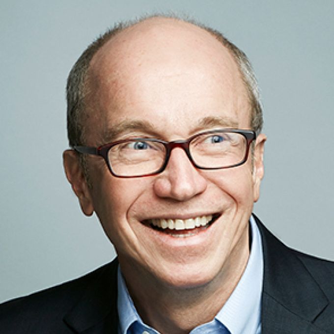 A man smiles brightly in trendy eyeglasses wearing a gray jacket and light blue shirt