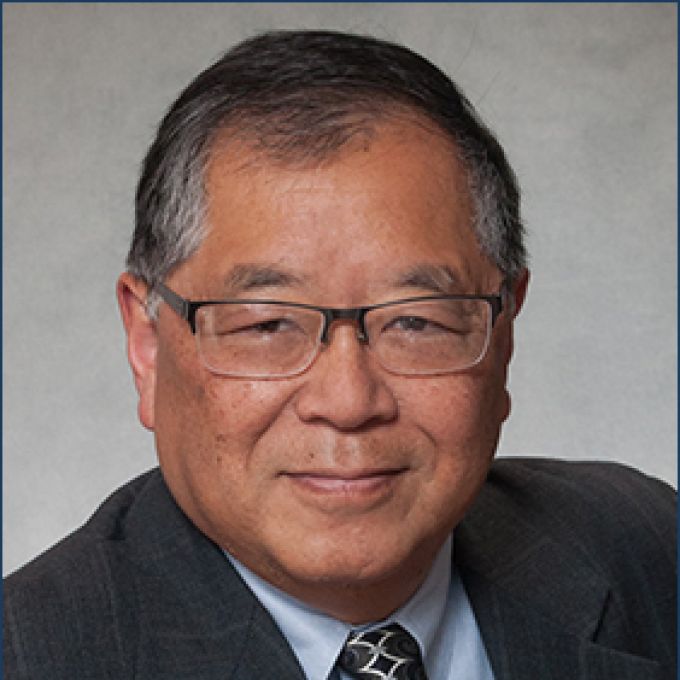 A man with graying hair and square-rim glasses poses in a dark jacket, light blue dress shirt, and patterned tie