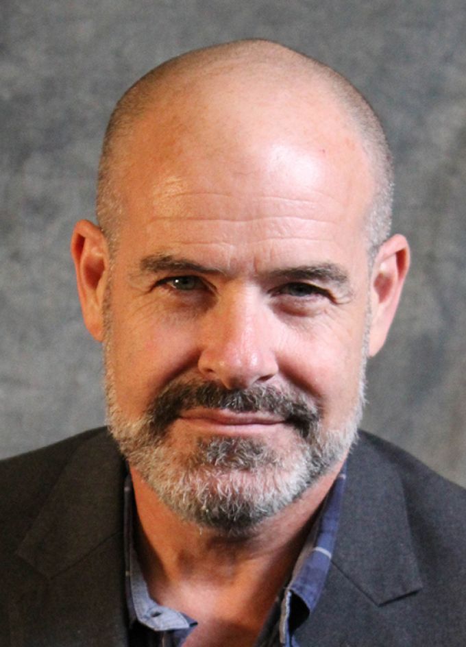 A bald man with neatly trimmed goatee poses in a gray jacket in front of a studio backdrop