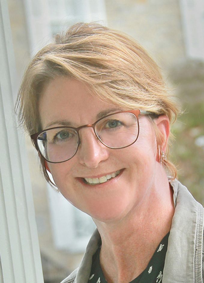 A woman with short blonde hair smiles in a tan jacket and glasses in front of a window