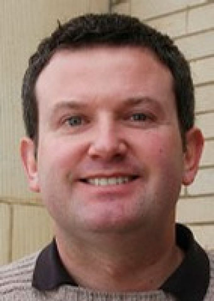 A man with short brown hair wearing a tan sweater and brown shirt smiles in front of a tan brick wall