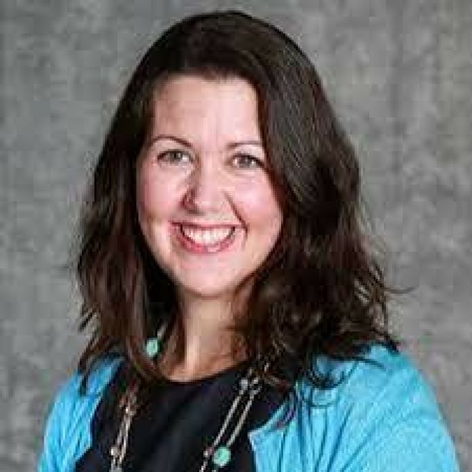 A woman with long brown hair smiles brightly in a turquoise jacket and black shirt