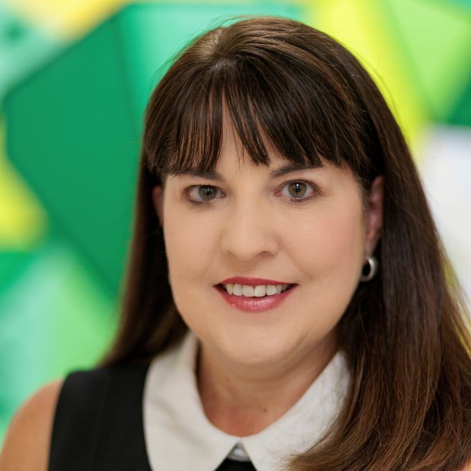 A woman with long brown hair poses in front of an abstract green and yellow background in a black jacket with gold buttons and a white collared shirt