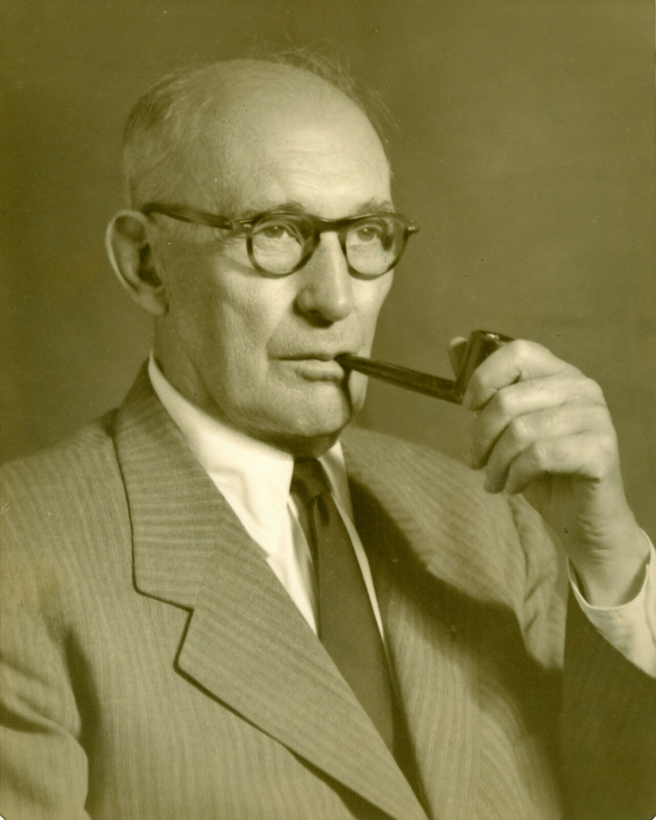 Duotone image of a bald man with tortise shell glasses and a pipe wearing a striped suit, dress shirt, and tie.