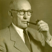 Duotone image of a bald man with tortise shell glasses and a pipe wearing a striped suit, dress shirt, and tie.