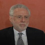 A man with gray hair and neatly trimmed beard poses in a dark jacket, white shirt, and striped tie in front of a terra cotta colored wall