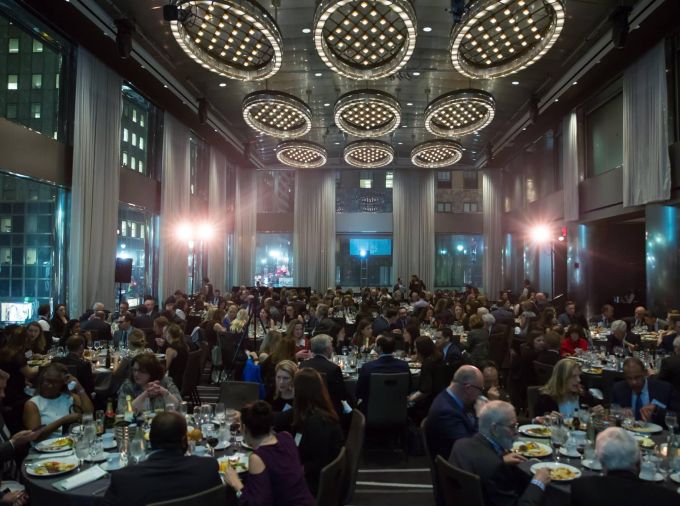 A grand ballroom with city skyscrapers lit up at night in the background with many people around banquet tables