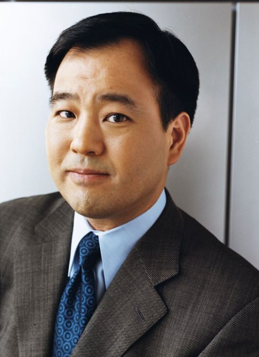 A man with black hair poses in a dark gray jacket, blue dress shirt, and dark blue tie.