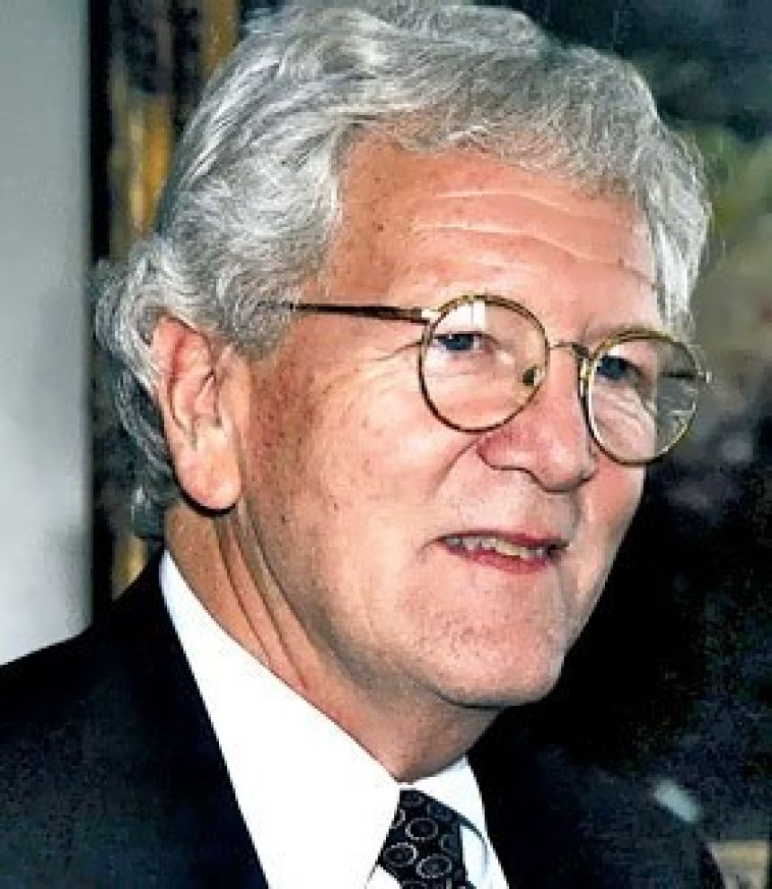 A man with gray hair and rounded glasses poses in dark jacket, white shirt, and tie